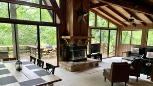 A cozy cabin interior with a stone fireplace, wooden furniture, and large windows offering a view of the outdoors.