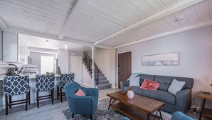 A modern living room with a gray sofa, blue chairs, and a wooden coffee table. A staircase leads to another room, and a kitchen area is visible in the background. - The Pineapple Suite, Whitewater Views steps to Sand  (San Clemente)