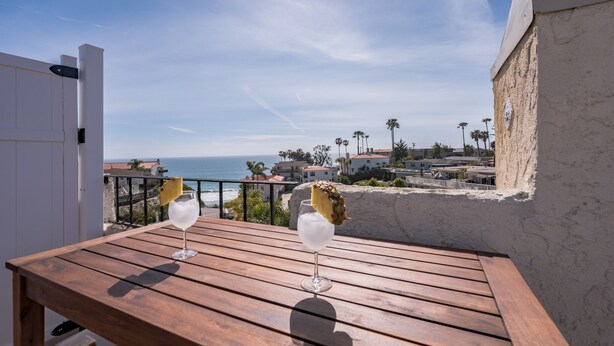 A wooden table with two glasses of a clear beverage, overlooking a coastal town with palm trees and houses. - The Pineapple Suite, Whitewater Views steps to Sand  (San Clemente)