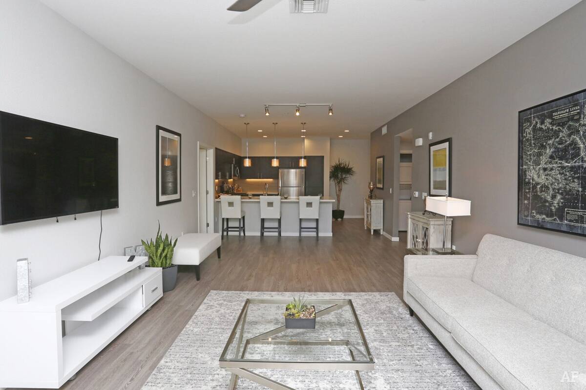 A modern living room with a flat-screen TV, a glass coffee table, a white sofa, and a kitchen in the background.