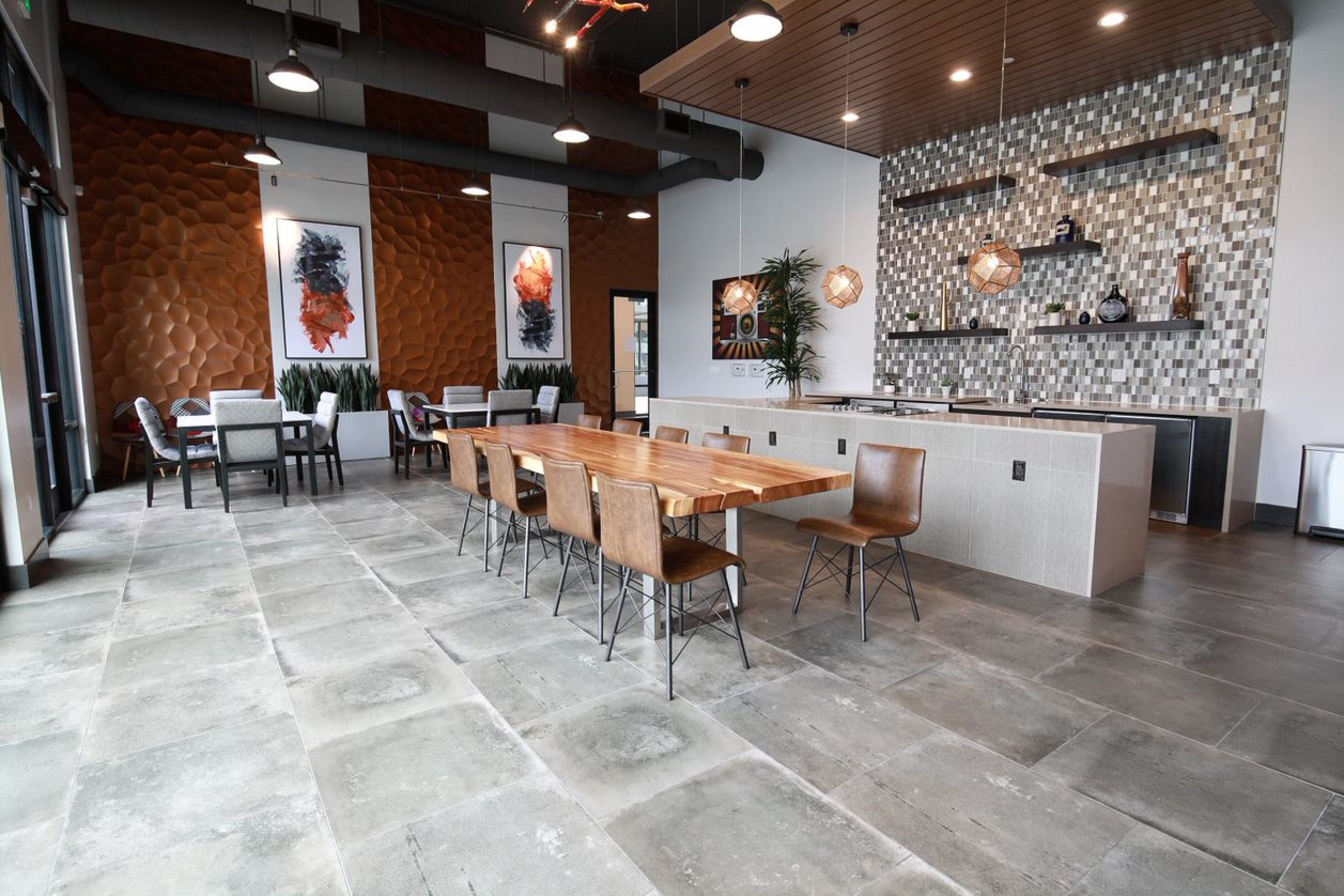 A modern dining area with a long wooden table and chairs, a kitchen area with a tiled backsplash, and a stone floor.