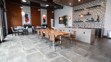 A modern dining area with a long wooden table and chairs, a kitchen area with a tiled backsplash, and a stone floor.