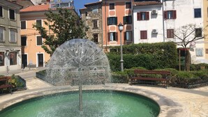 Ein Brunnen auf einem Platz, umgeben von Gebäuden mit Balkonen und Fensterläden.