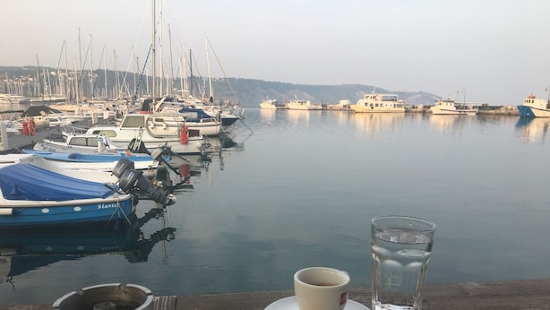 Ein Yachthafen mit vertäuten Booten, ein Holztisch mit einer Kaffeetasse, einem Glas Wasser und einer Zigarettenpackung.