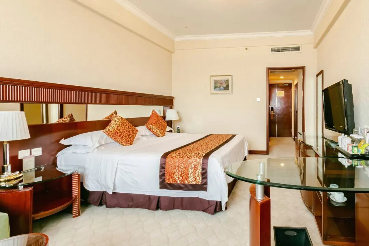 A hotel room with a bed, bedside tables, a glass coffee table, a TV, and a door leading to another room.