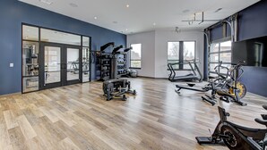 Ein großzügiger Fitnessraum mit unterschiedlichen Trainingsgeräten, großen Fenstern und Holzboden.