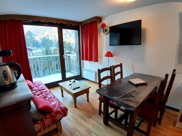 A cozy room with a sofa, a small table, a dining table with chairs, a TV mounted on the wall, and a balcony with red curtains.