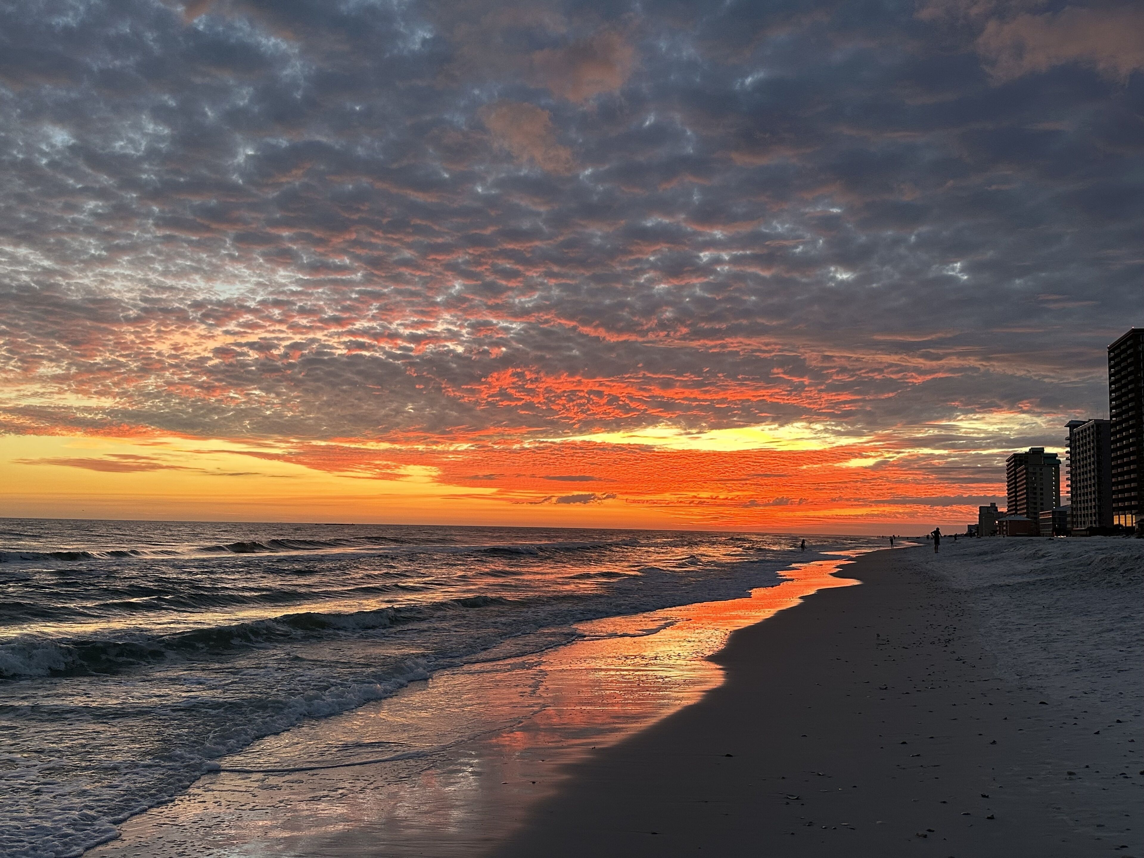 A beach with a sunset, waves, and buildings in the background.