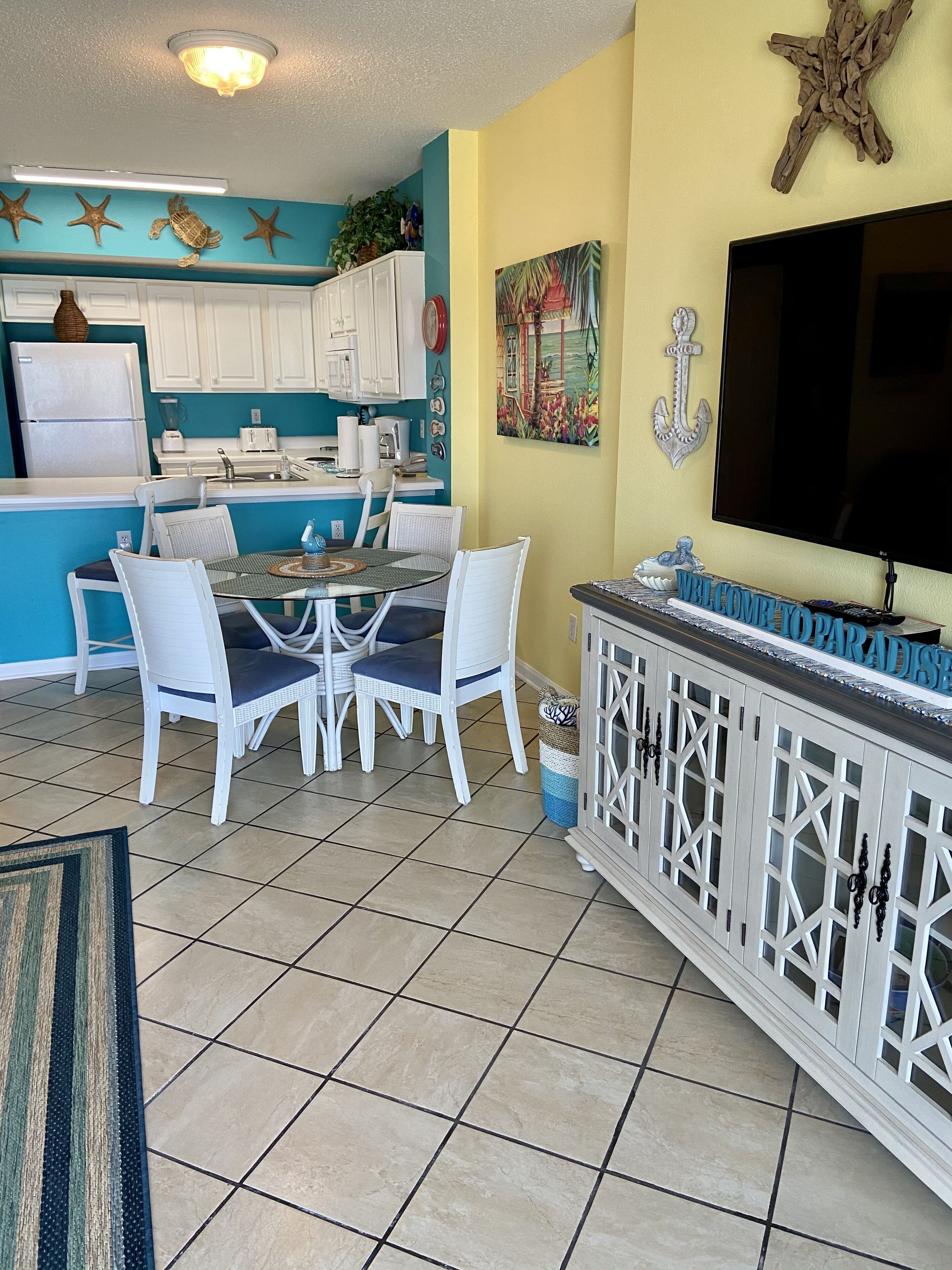 Cocina compacta con armarios blancos, comedor con sillas blancas y zona de bar con televisor.