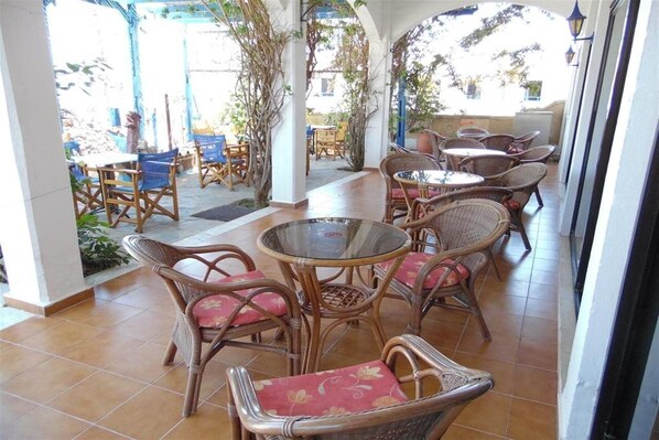 An outdoor patio area with wicker furniture and glass-top tables, surrounded by plants and a covered walkway. - Panorama Gennadi (Rhodes)