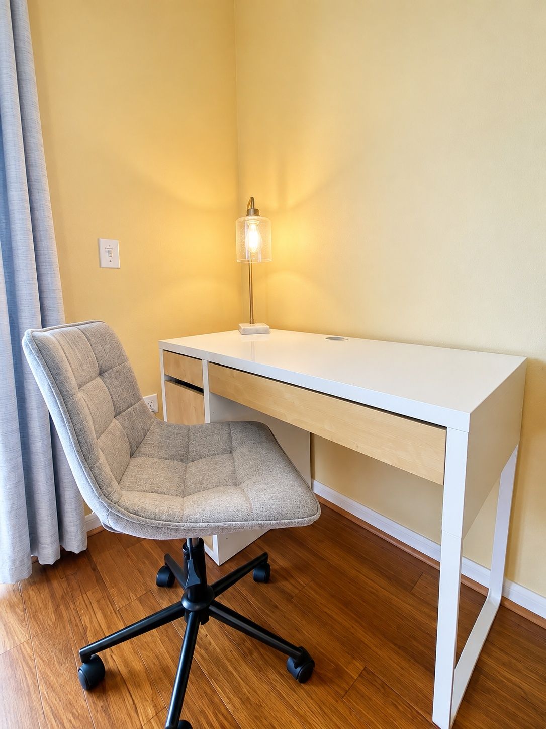Un espacio de trabajo con un escritorio blanco, una silla con acolchado gris y una lámpara de pie.