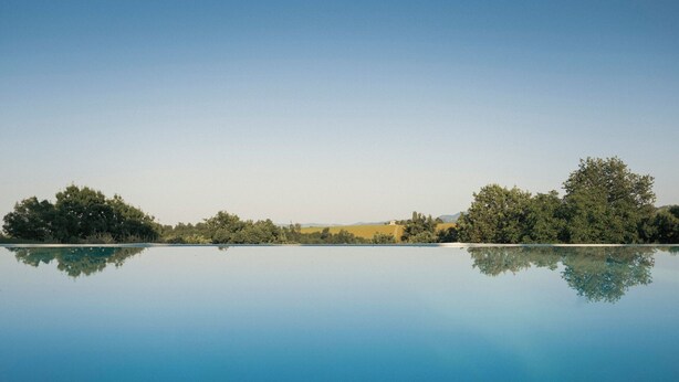 Uma piscina ao ar livre tranquila, com águas claras que refletem as árvores ao redor e um edifício distante.