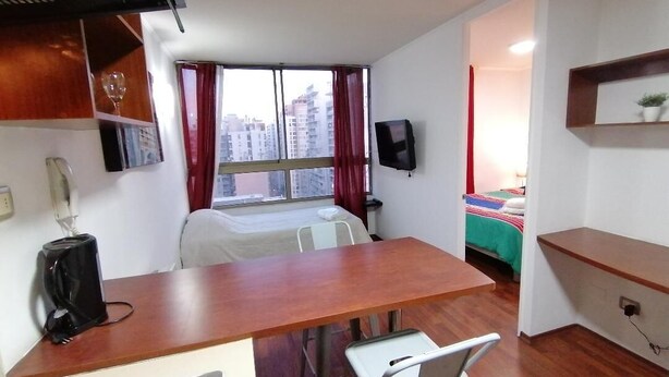 A compact room with a kitchenette, a bed, a TV, and a window with a city view. - Santiago Moneda Suites 1802 (Santiago)