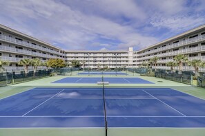 Una cancha de tenis con una red en el centro, rodeada por una valla, y un edificio con varios balcones al fondo.