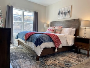 Une chambre à coucher avec un grand lit, une table de chevet, une lampe, une fenêtre avec des rideaux et un tableau au mur.