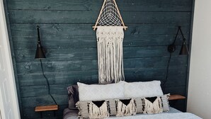 Una cama con una colcha blanca y dos almohadas, un tapiz macramé sobre la cama y dos lámparas de noche sobre una pared de madera oscura.