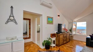 Un vaste espace de vie doté d’un parquet en bois disposé en chevrons, d’une grande fenêtre offrant une vue sur la mer, d’un téléviseur et d’une horloge décorative en forme de tour Eiffel.