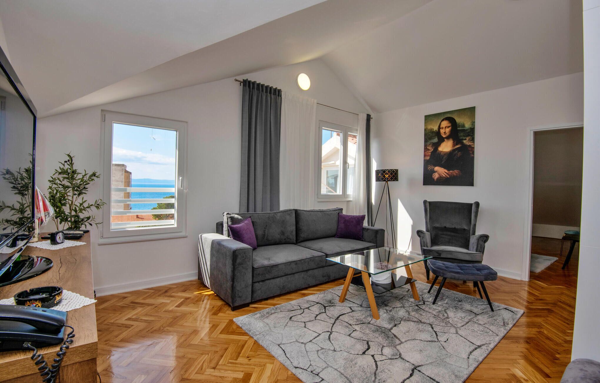 Ein modernes Wohnzimmer mit grauem Sofa, einem Glastisch und einem Sessel. An der Wand hängt ein Gemälde der Mona Lisa, und es gibt einen Balkon mit Blick sowie Holzdielenboden.
