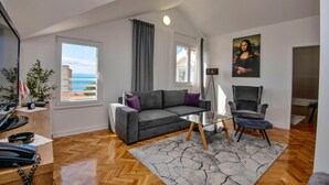 Un salon moderne avec un canapé gris, une table basse en verre et un fauteuil. Un tableau représentant la Joconde est accroché au mur, il y a un balcon avec vue et un plancher en bois.