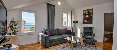 A modern living room with a grey sofa, a glass coffee table, and a chair. There is a painting of the Mona Lisa on the wall, a balcony with a view, and a wooden floor.