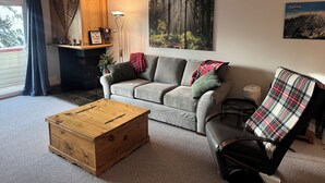 A living room with a grey sofa, a wooden coffee table, a plaid blanket, and a painting of a forest. - Big White Ski-In Ski-Out at Ponderosa in Village (Big white)