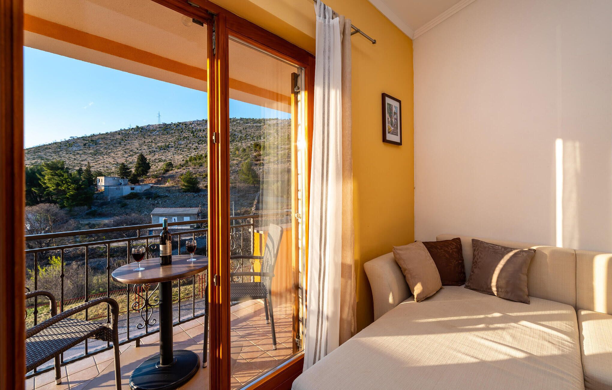 Ein Zimmer mit Balkon, einer Couch, einem Tisch mit Weingläsern und einer Flasche sowie Blick auf eine hügelige Landschaft.