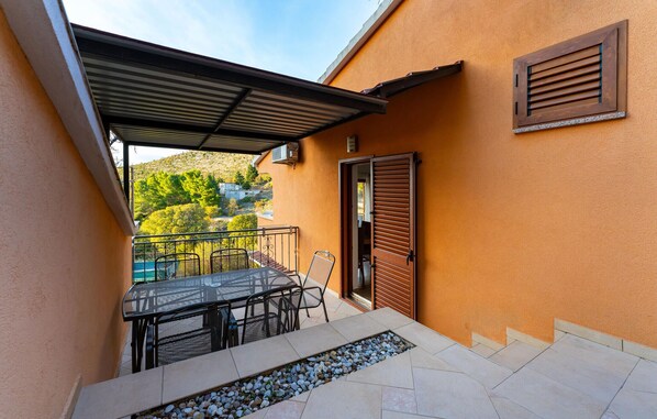 A covered outdoor seating area with a table and chairs, a door, and a window with shutters.