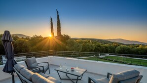 A terrace with outdoor furniture, a table, and a view of the sunset over a landscape with trees and hills.