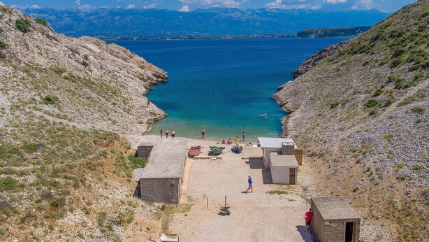 Una zona costera con una playa rocosa, un pequeño muelle y una vista despejada del mar y montañas a lo lejos.