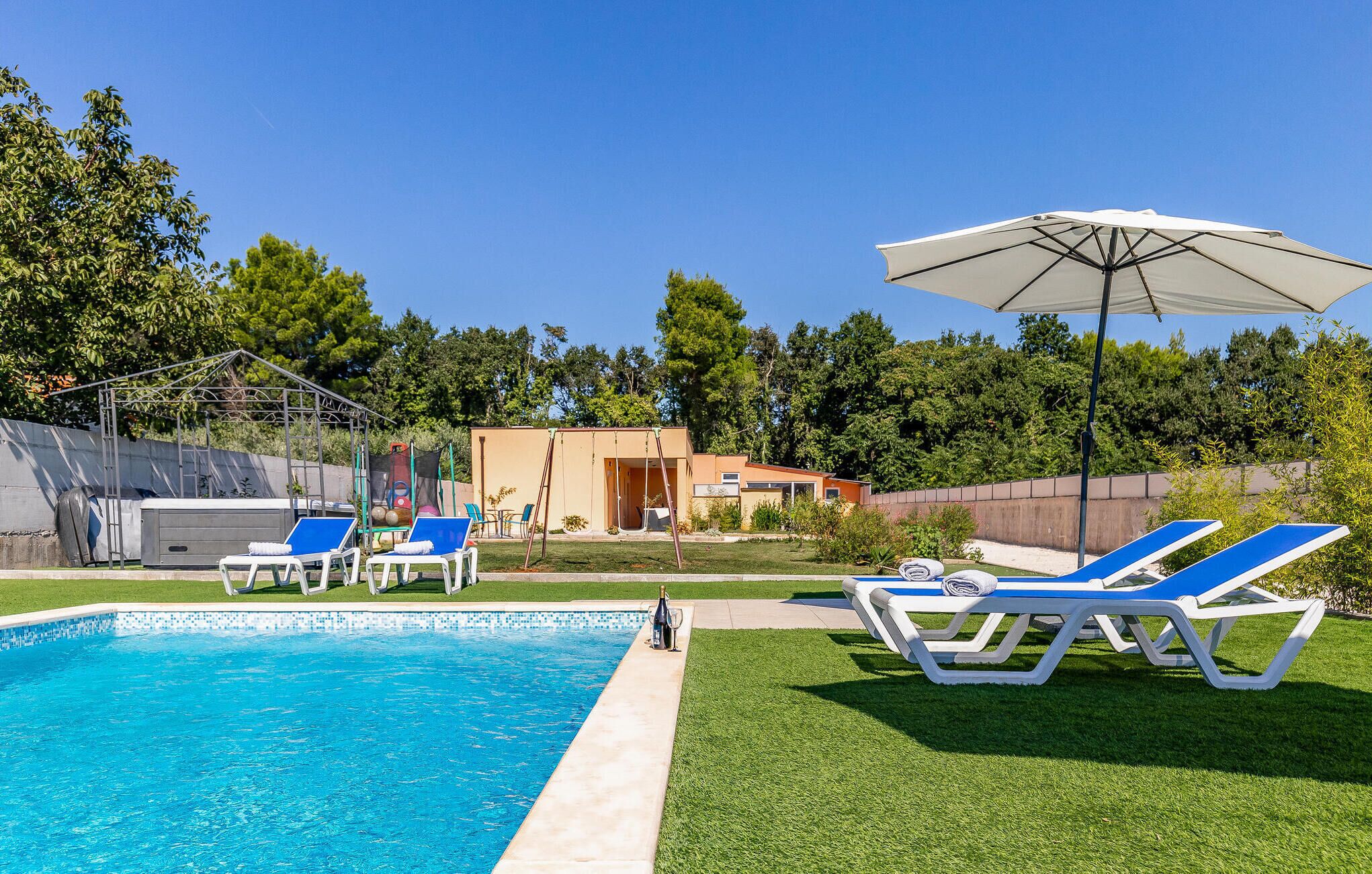 A swimming pool with lounge chairs and an umbrella, surrounded by a grassy area and trees.
