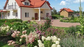 Una casa de dos pisos con techo de tejas rojas, un balcón y un jardín con diversas flores y plantas.