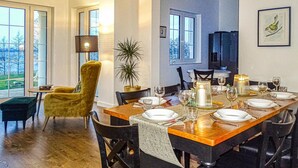 Un comedor con una larga mesa de madera dispuesta para seis personas, rodeada de sillas negras. Hay dos plantas en macetas, una lámpara y un cuadro en la pared.