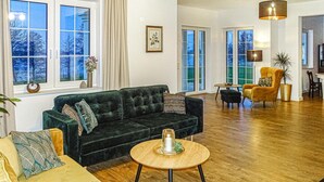 Un salón amplio con un sofá verde oscuro, una mesa de centro redonda de madera y un sillón amarillo. Se observan ventanas grandes con cortinas, un cuadro enmarcado en la pared y una lámpara colgante.