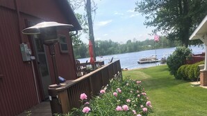 Une pelouse bien entretenue, bordée d’une rangée de fleurs roses, une terrasse en bois et une vue sur un lac où naviguent des bateaux.