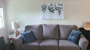A living room with a grey sofa, two blue cushions, a wooden side table with a lamp, and a wall-mounted painting of vases with flowers. - The Corner Shed- Norris Lake or the Mountains (Jacksboro)