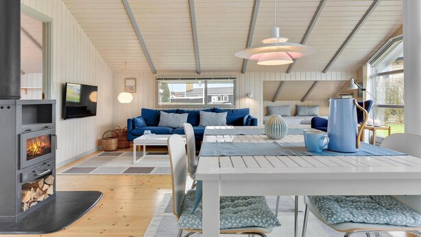A cozy living room with a fireplace, a blue sofa, a dining table, and a TV mounted on the wall. - Lovely home in Broager with WiFi (Broager)