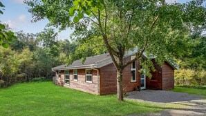 Een klein houten huisje met een schuin dak, omgeven door weelderige begroeiing en een verzorgd gazon.