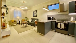 A modern living room with a sofa, coffee table, and a TV. A kitchen area with a stove and microwave is visible.