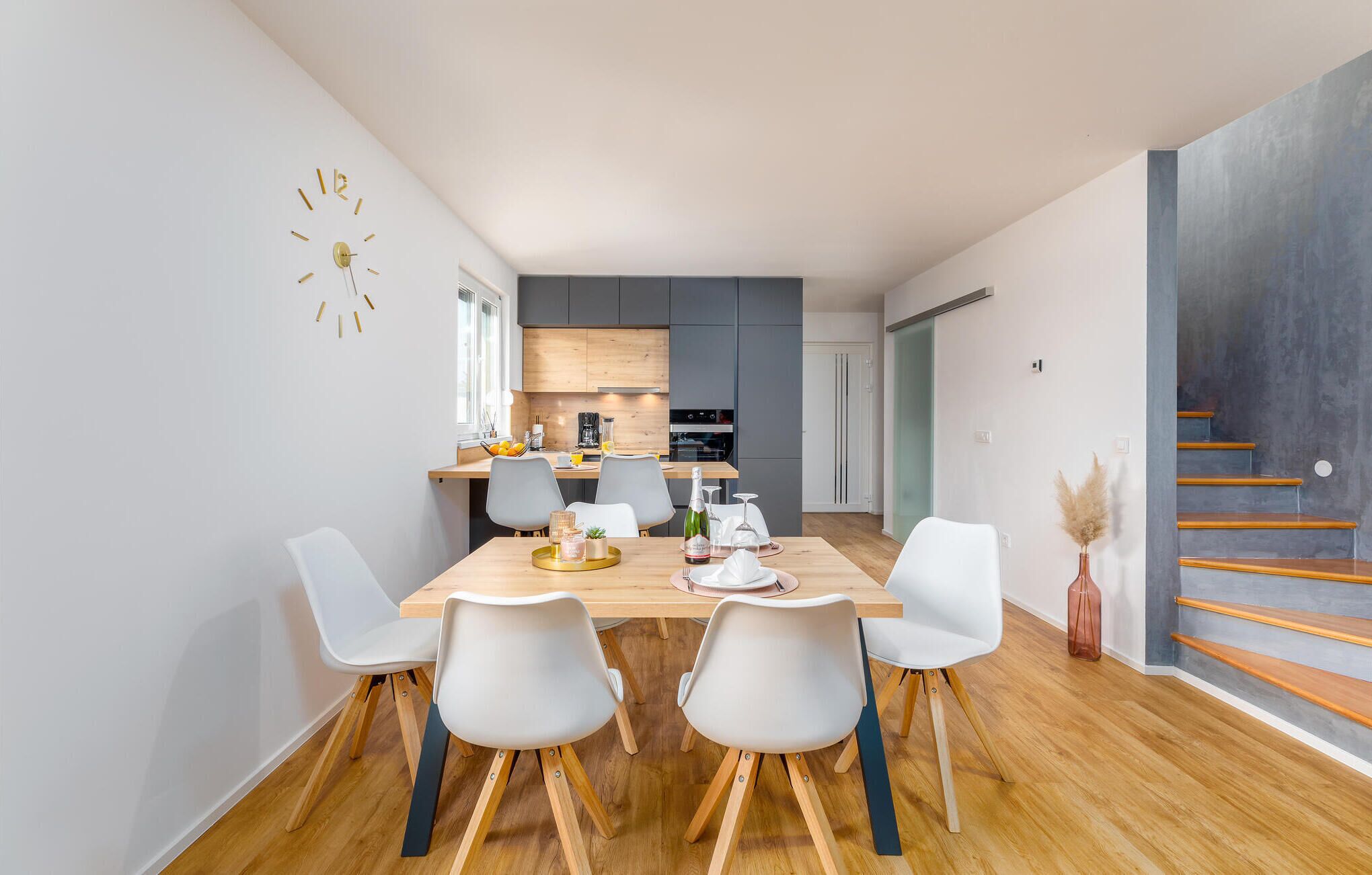 A modern kitchen with wooden cabinets, a dining area with a wooden table and white chairs, and a staircase with wooden steps.