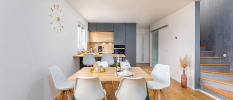 A modern kitchen with wooden cabinets, a dining area with a wooden table and white chairs, and a staircase with wooden steps.