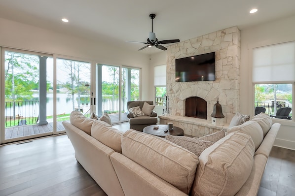 A spacious living room with a stone fireplace, a ceiling fan, and a large window overlooking a deck.