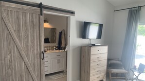 Una habitación con una puerta corredera que da a un baño, un aparador de madera con un televisor encima, una silla y una mochila en el suelo.