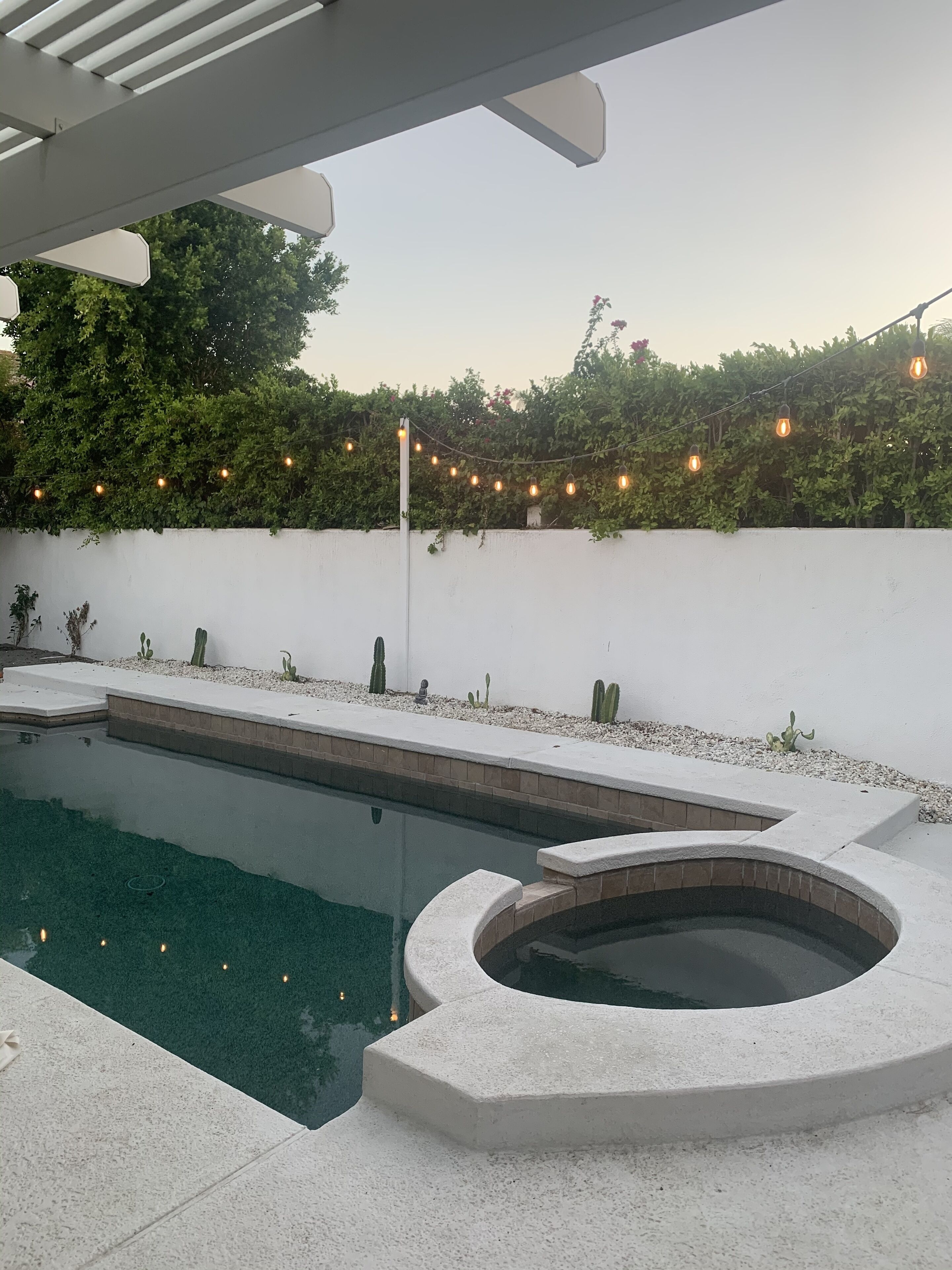 Un área de piscina moderna al aire libre con un jacuzzi, rodeada por un muro blanco y vegetación.
