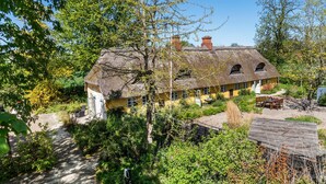A traditional thatched-roof house with a stone foundation, surrounded by lush greenery and a clear sky.