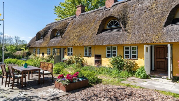 A traditional thatched-roof house with a yellow exterior, a wooden table with chairs, and a flower bed in the front yard.