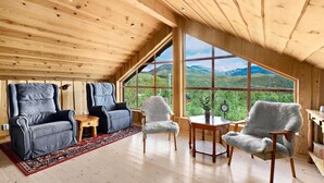 Interior acogedor de una cabaña con techos de madera, un ventanal con vistas a la montaña, dos sofás grises, una mesita de madera y un sillón blanco mullido.