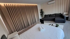 A modern hotel room with a large bed, a grey sofa, a small table, and a potted plant. - Figueira Beach Apartment (Buarcos)