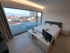A modern hotel room with a large balcony, a bed with neatly folded towels, a TV, and a small table.