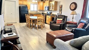 A living room with a dining table, leather chairs, a sofa, a coffee table, a TV stand, and a kitchen area with wooden cabinets and appliances. - Greater Zion Villa at Sports Village (St. George)
