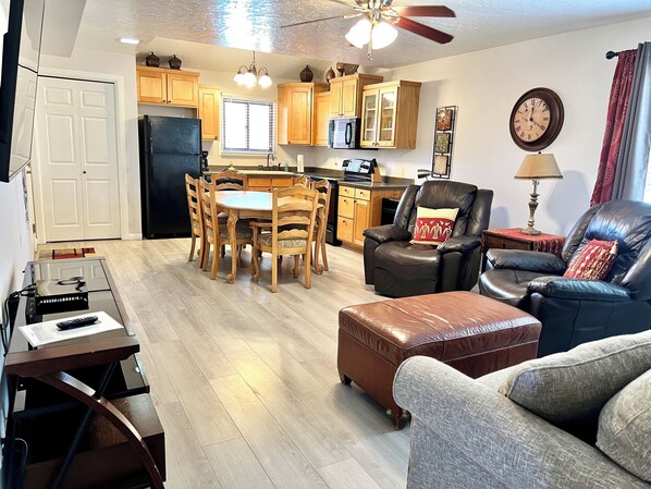 A living room with a dining table, leather chairs, a sofa, a coffee table, a TV stand, and a kitchen area with wooden cabinets and appliances.